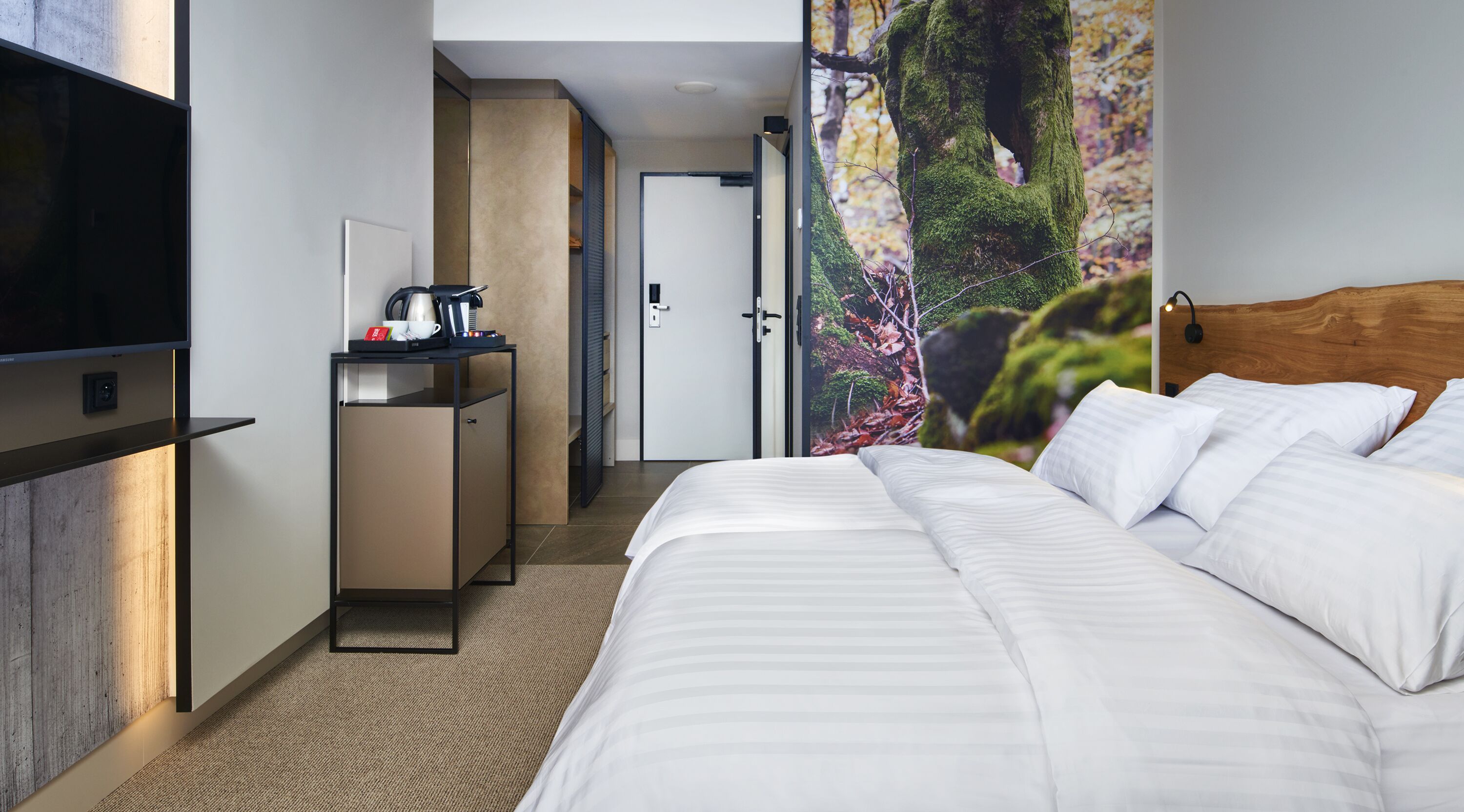 A hotel room with a bed, a desk with a coffee maker, a TV, and a door leading to another room.