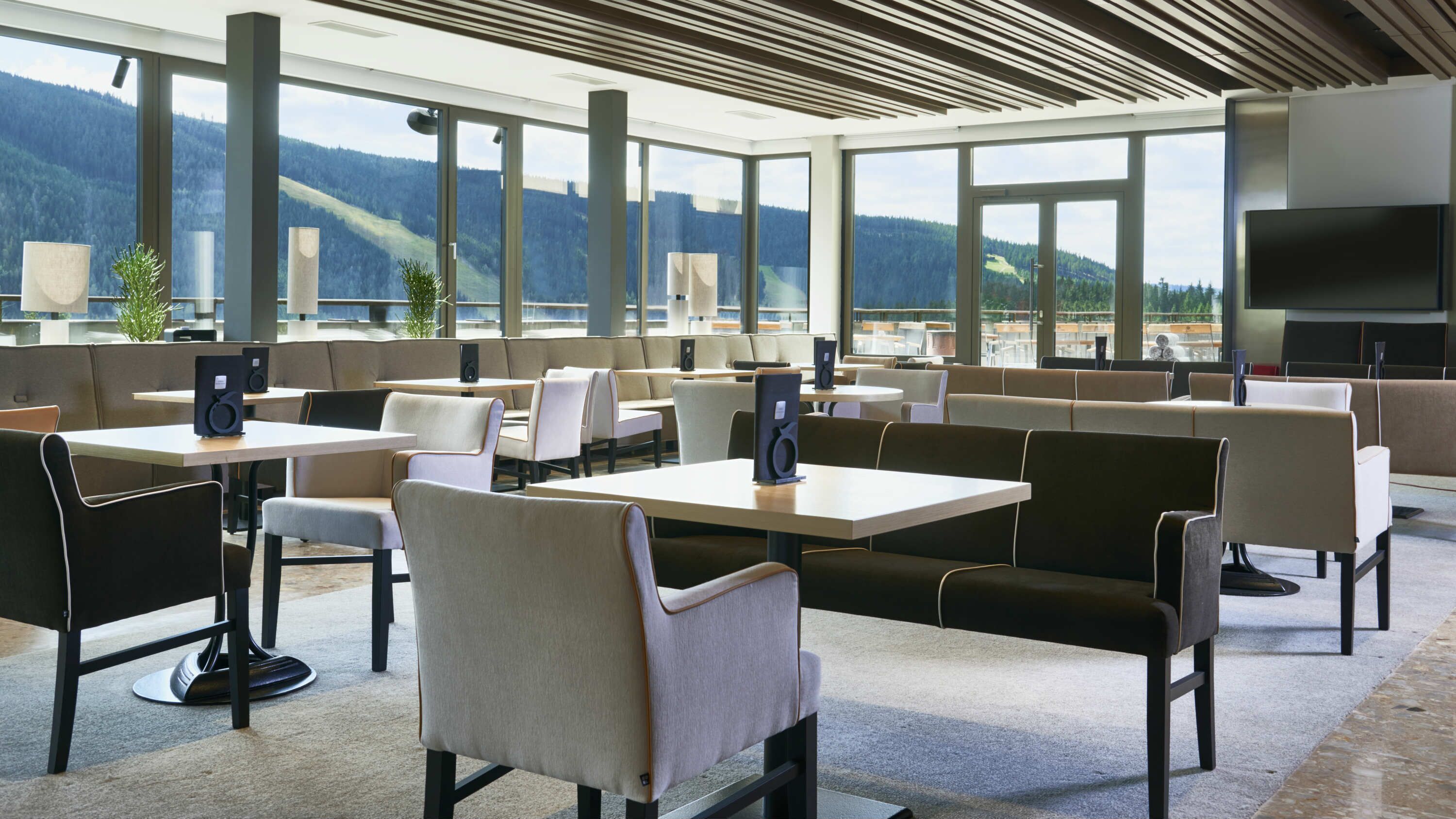 Un restaurant moderne avec de hauts plafonds et de grandes fenêtres offrant une vue sur les montagnes. Les tables et les chaises sont disposées pour accueillir les clients.
