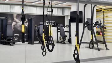 Un gimnasio con diversos equipos de ejercicio, incluyendo entrenadores de suspensión TRX, máquinas de pesas y un muro con espejos.