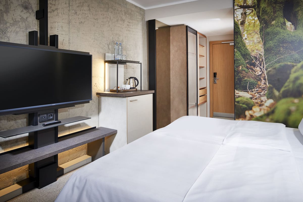 A modern hotel room with a large flat-screen TV, a wooden desk, a bed with white linens, and a forest-themed wall mural.