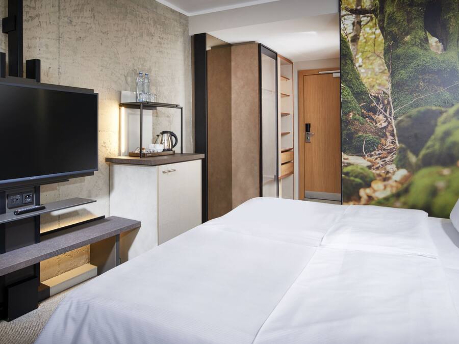 A modern hotel room with a large flat-screen TV, a wooden desk, a bed with white linens, and a forest-themed wall mural.