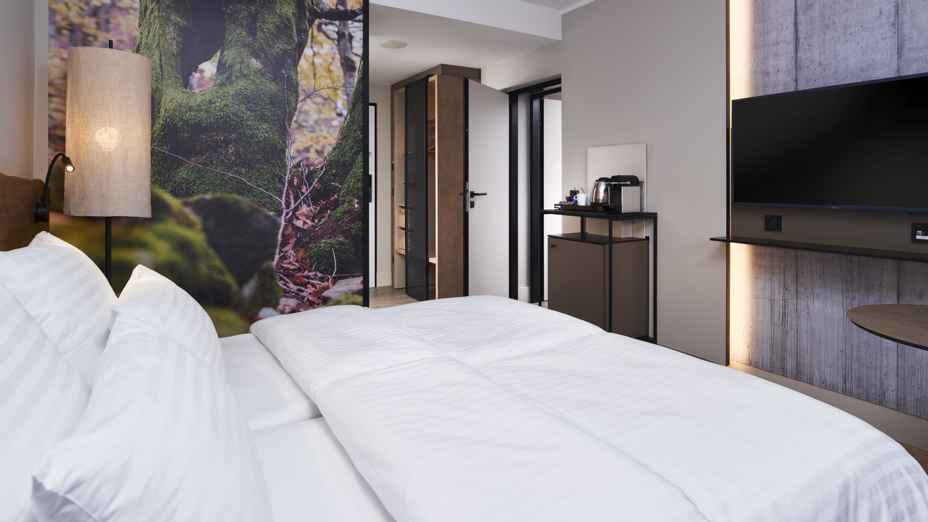 Una habitación de hotel moderna con una cama grande, un televisor montado en la pared y vistas a un bosque a través de un ventanal.