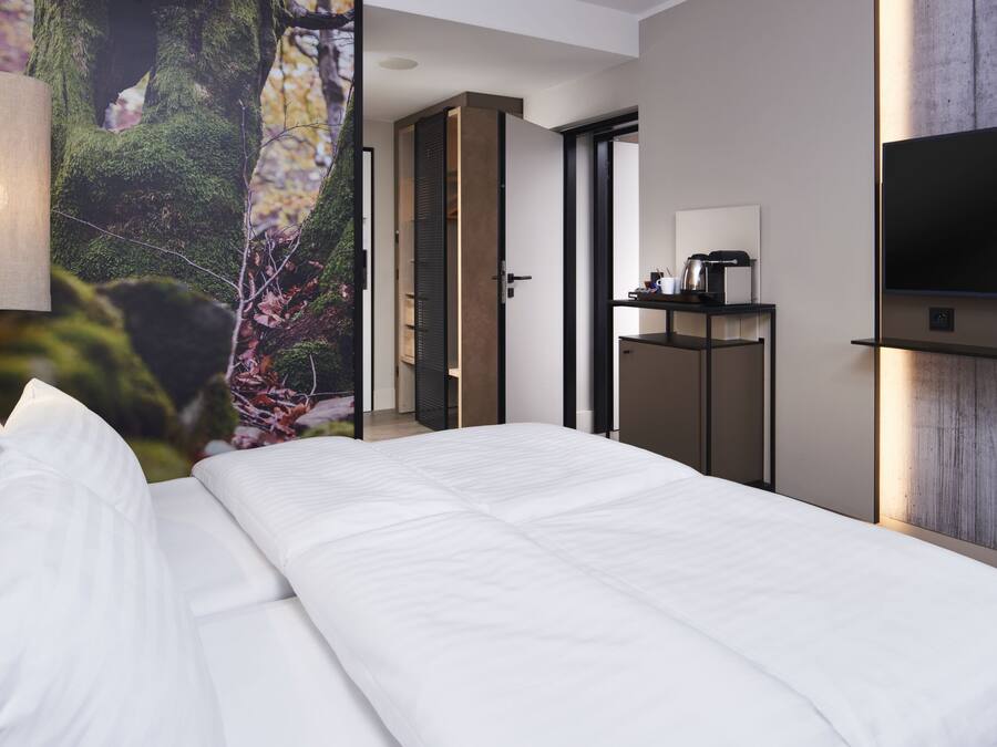 Una habitación de hotel moderna con una cama grande, un televisor montado en la pared y vistas a un bosque a través de un ventanal.
