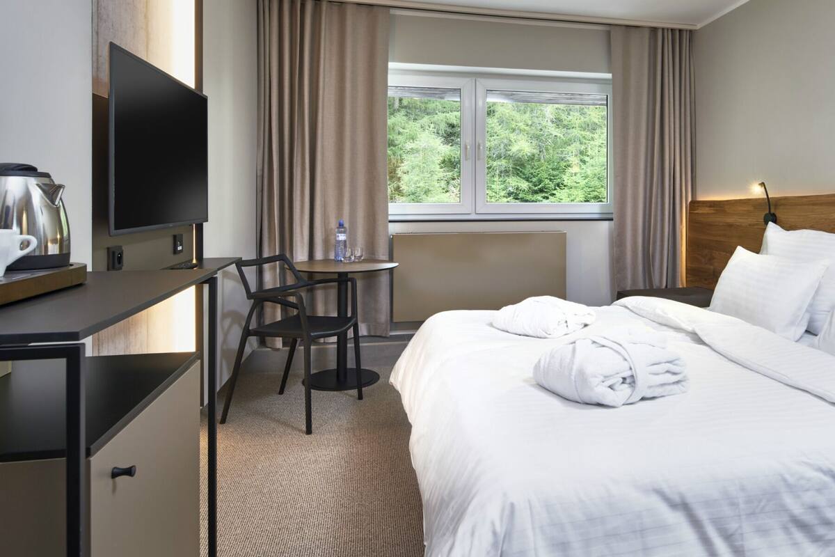 A hotel room with a bed, a desk with a TV, a chair, a window with curtains, and a nightstand with a lamp.