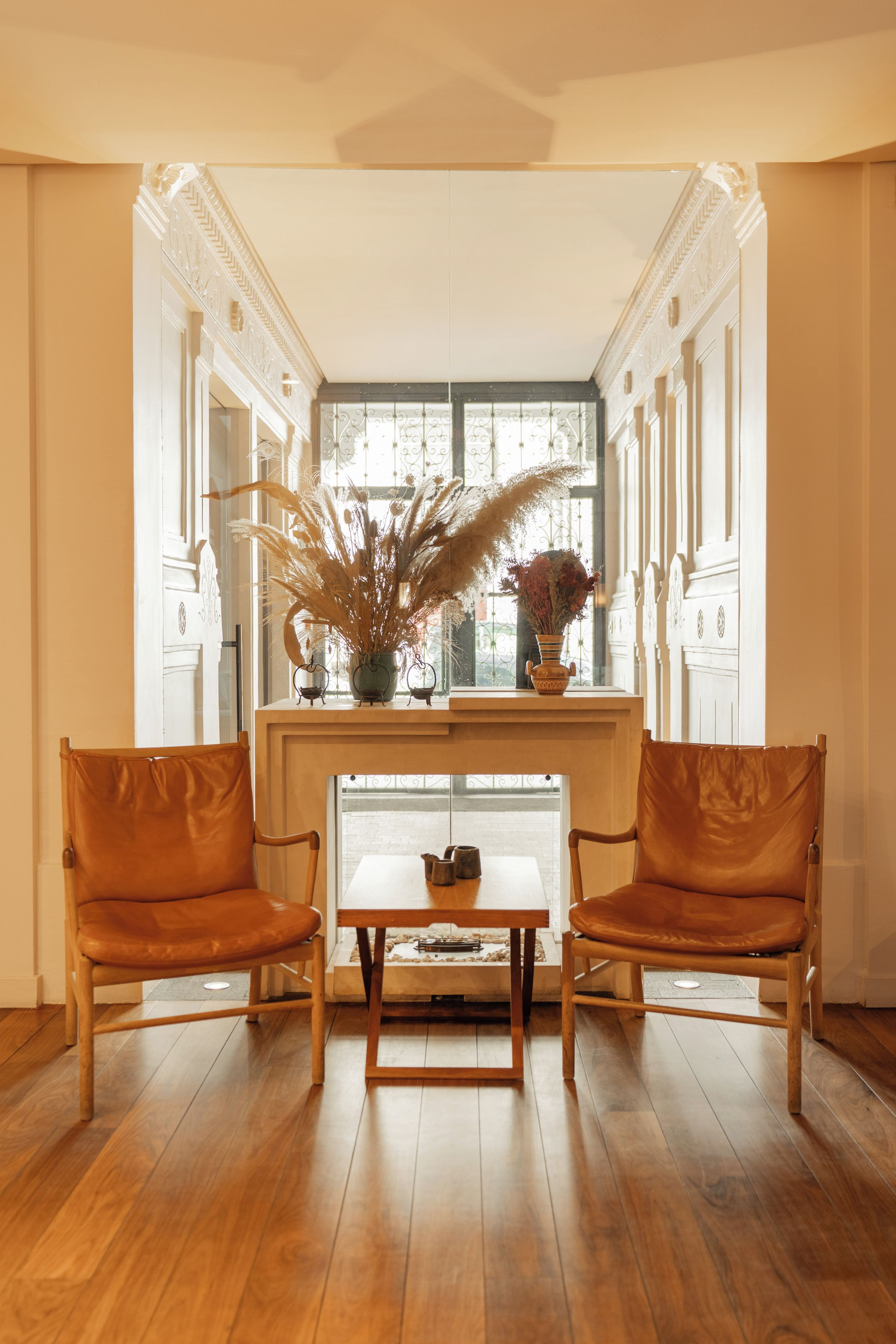 Un coin repas comprenant deux fauteuils en cuir, une table en bois et un vase avec des plantes séchées, posé sur une console.