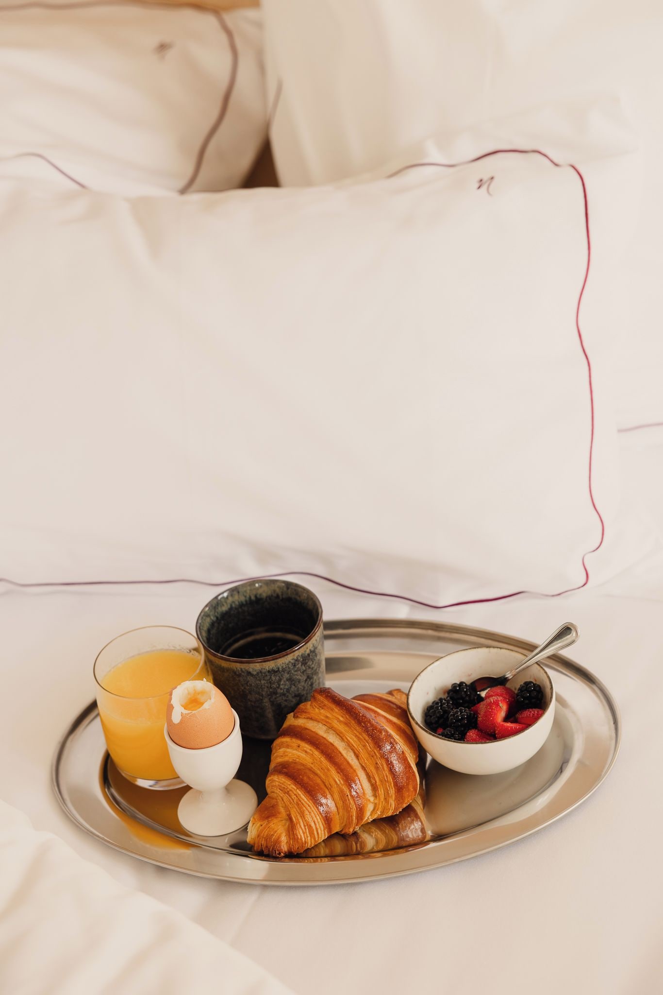 Un safata de petitdejuni amb suc d'aranja, un croissant, un bol de baies, una tassa de cafè i un gotet de iogurt sobre un llit amb roba blanca i almoades.
