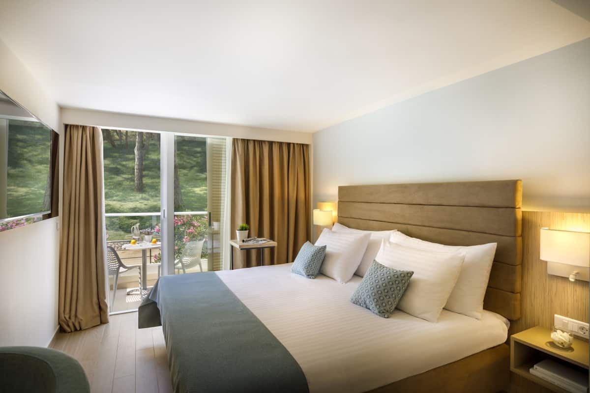 A hotel room with a large bed, a nightstand, a lamp, and a view of a balcony with outdoor furniture.