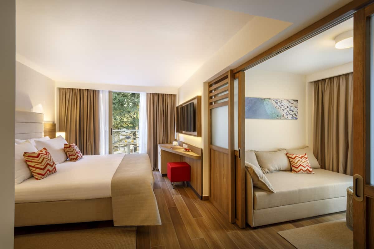 A modern hotel room with two beds, a desk, and a large window offering a view of greenery.