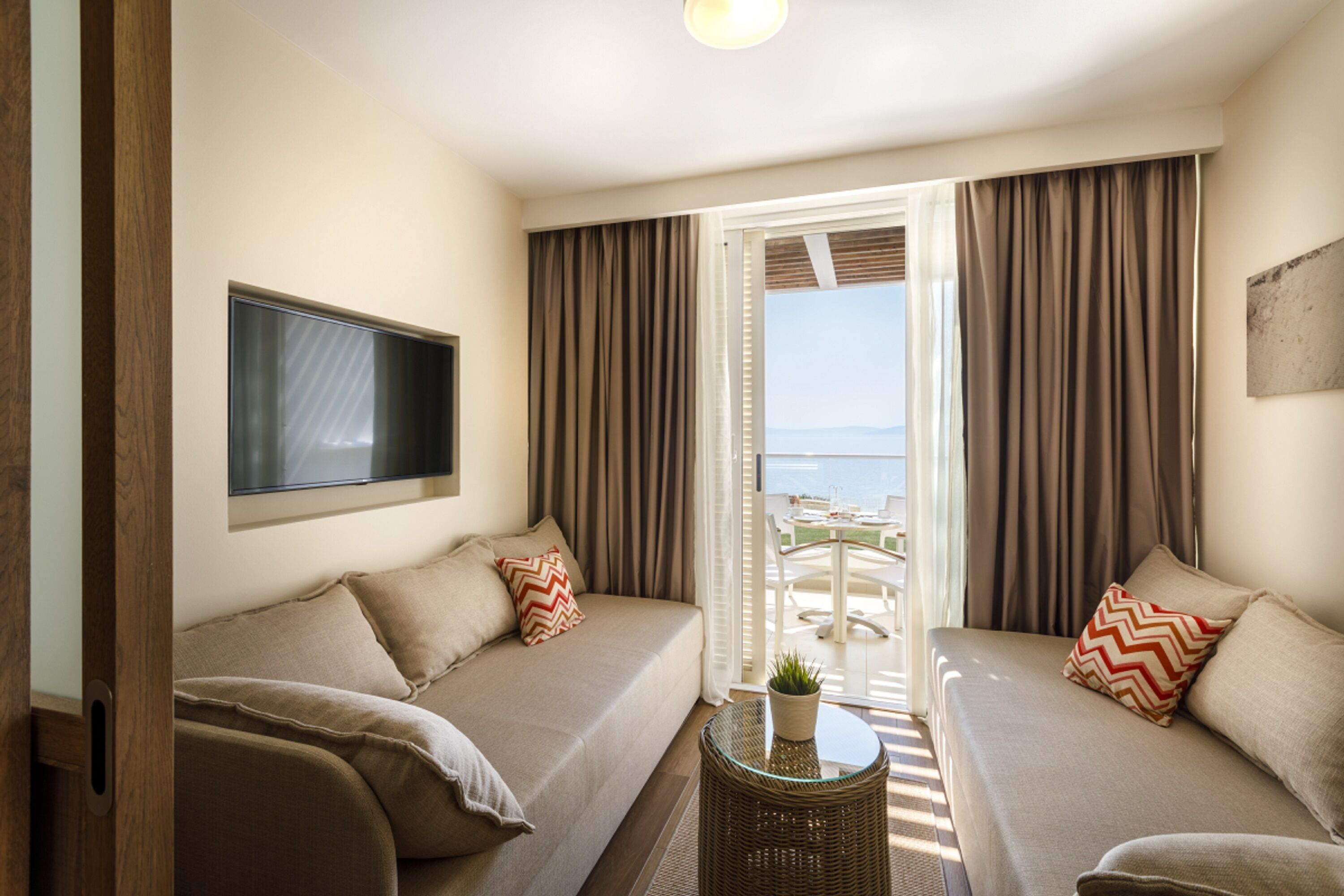 A modern hotel room with a sofa set, a flat-screen TV, and a balcony with a view of the sea.
