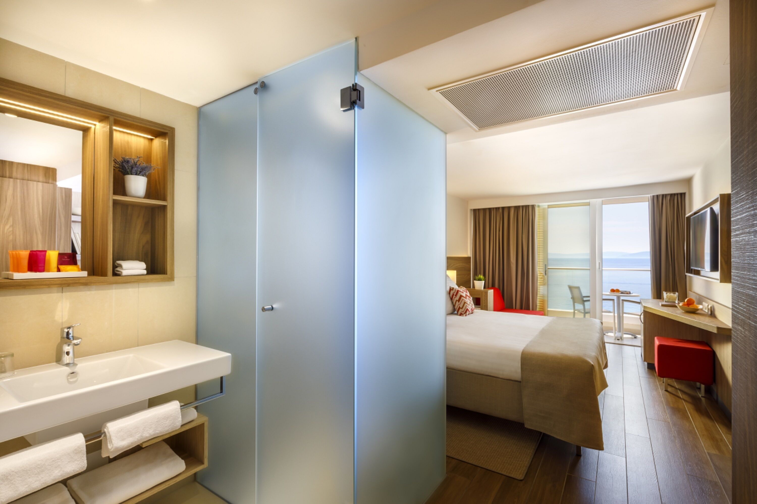 A hotel room with a large bed, a bathroom with a sink and mirror, a small table with a chair, and a balcony with a view of the ocean.