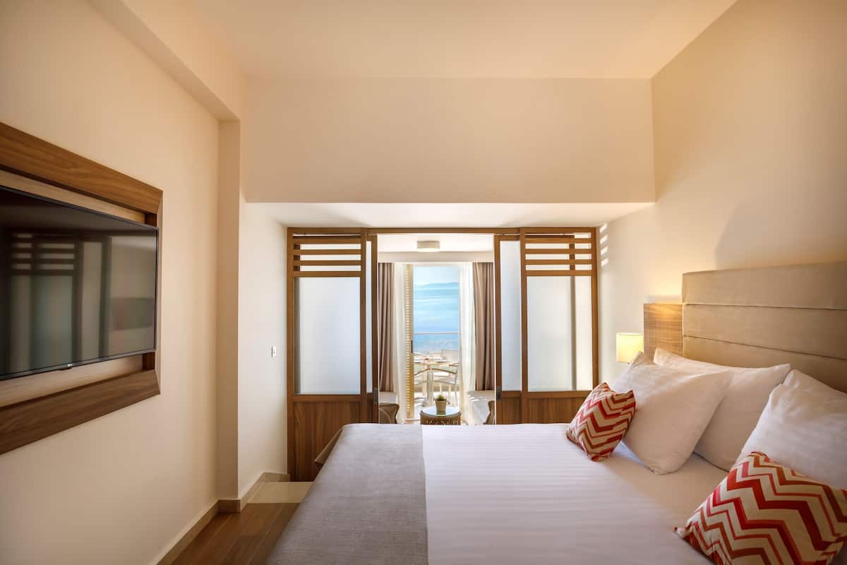 A hotel room with a bed, a TV mounted on the wall, and a view of the sea through a large window.
