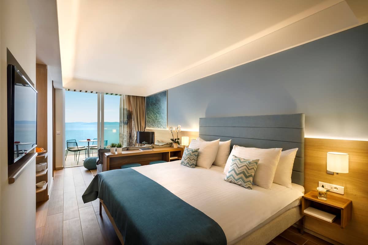 A hotel room with a large bed, a desk with a computer, a TV, and a view of the sea.