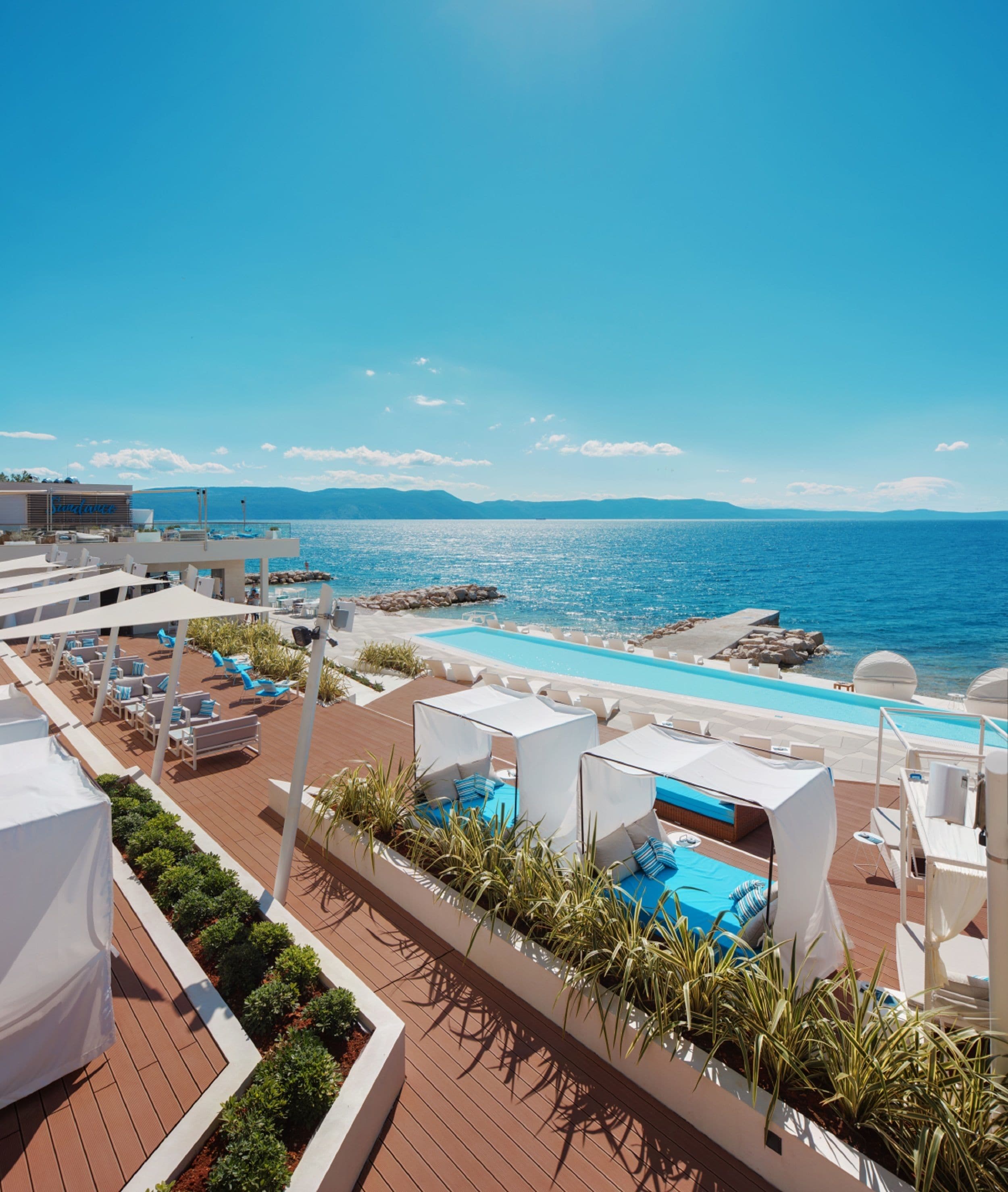 Uma área de piscina no terraço com espreguiçadeiras e guarda-sóis, com vista para o mar azul e límpido.