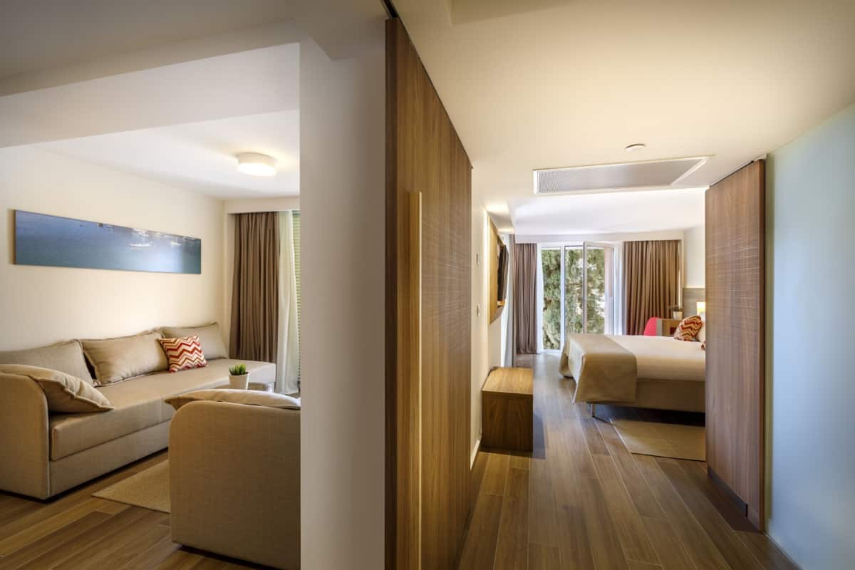 A modern hotel room with a sofa, a bed, a wooden floor, and a view of greenery outside.