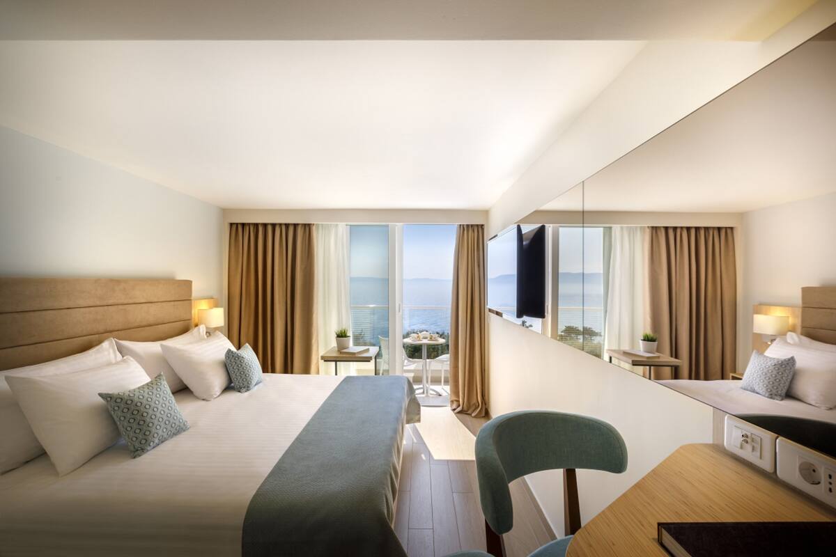 A hotel room with a large bed, a desk, a chair, a TV, and a balcony with a view of the ocean.