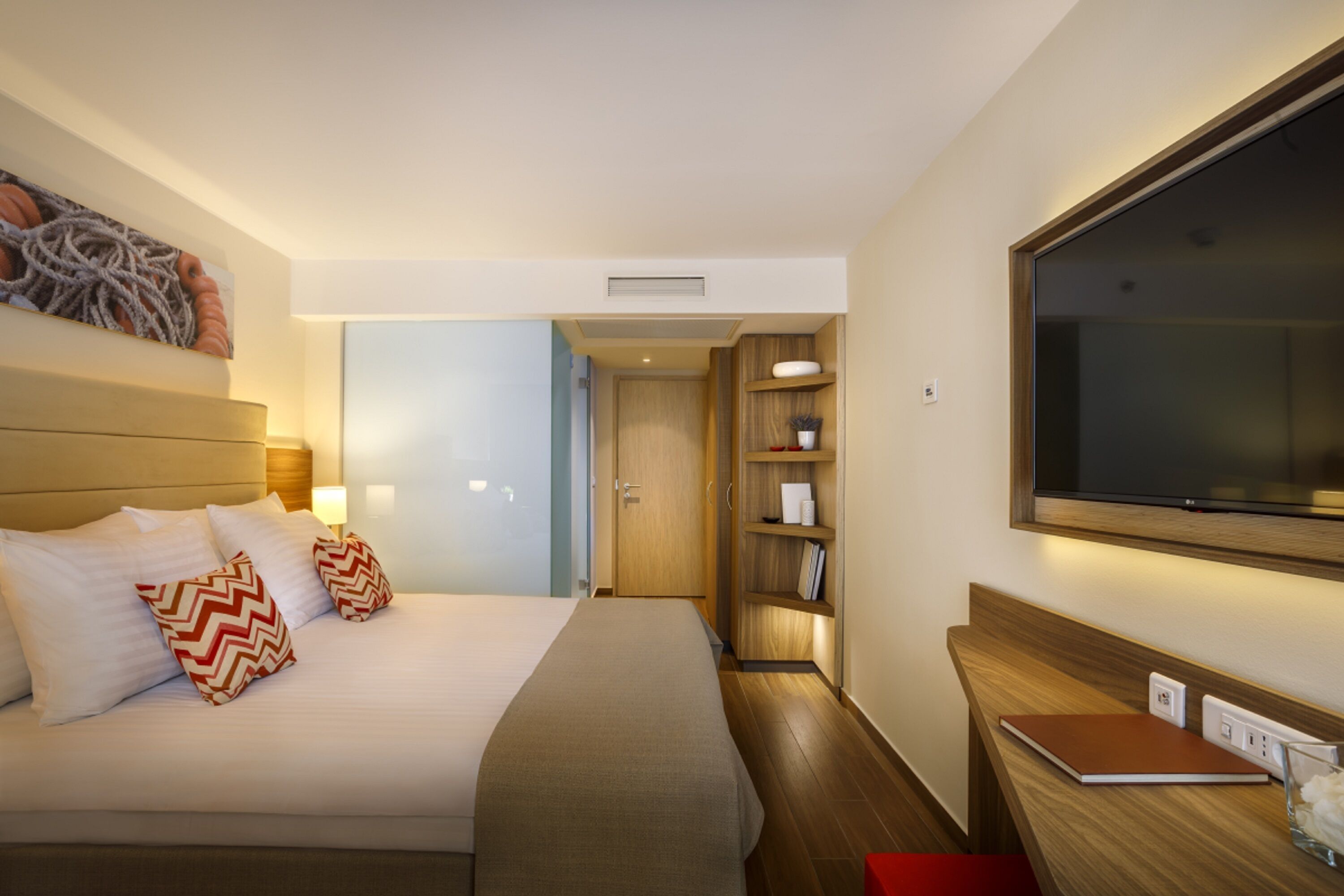 A hotel room with a large bed, a TV mounted on the wall, a wooden side table, and a bookshelf.