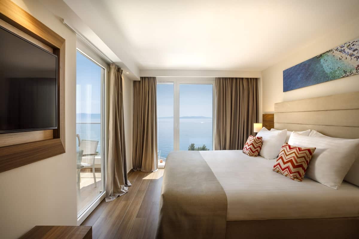 A hotel room with a large bed, a TV, a balcony with a view of the sea, and a painting on the wall.
