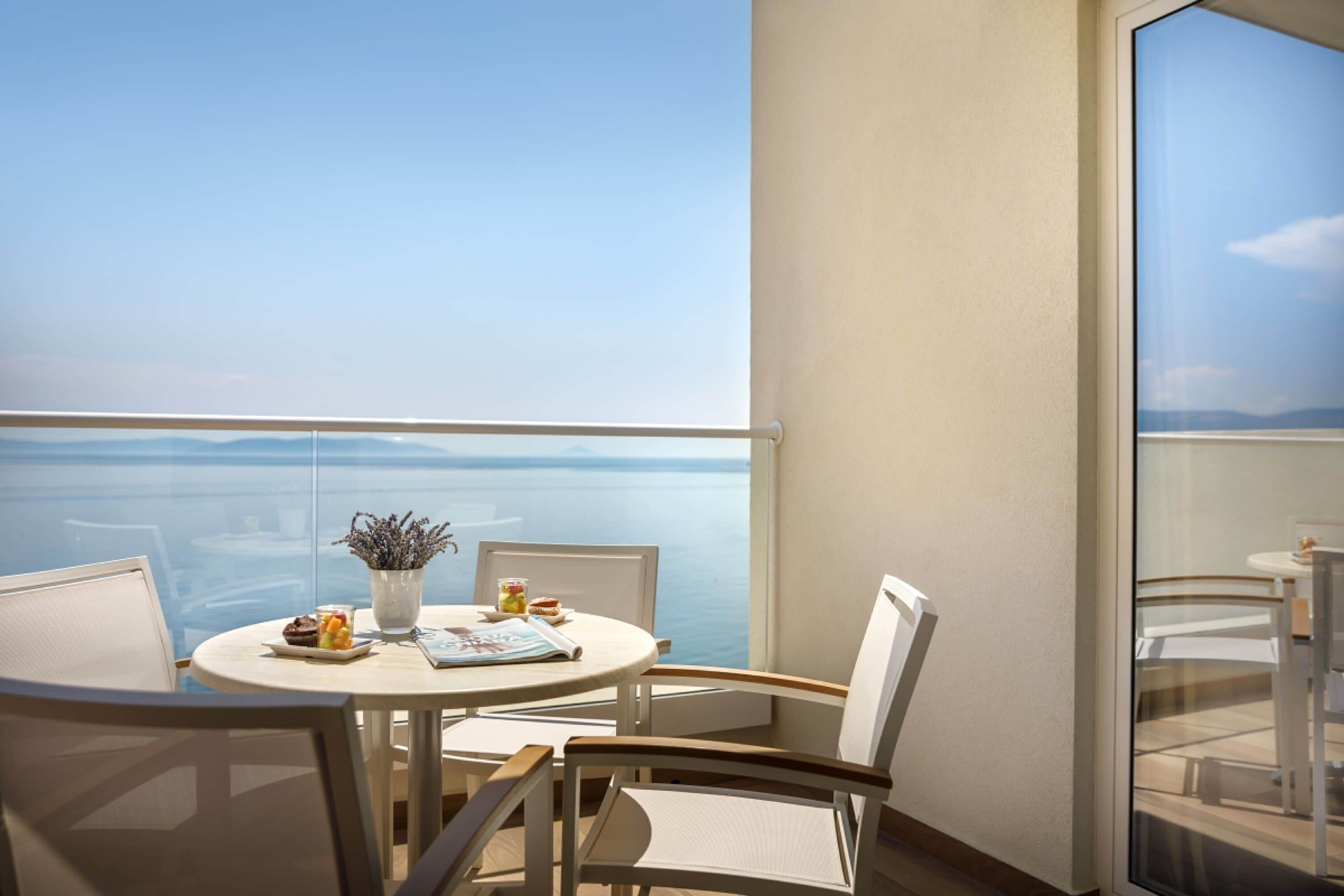 A balcony with a round table, chairs, a vase with flowers, and a view of the sea.