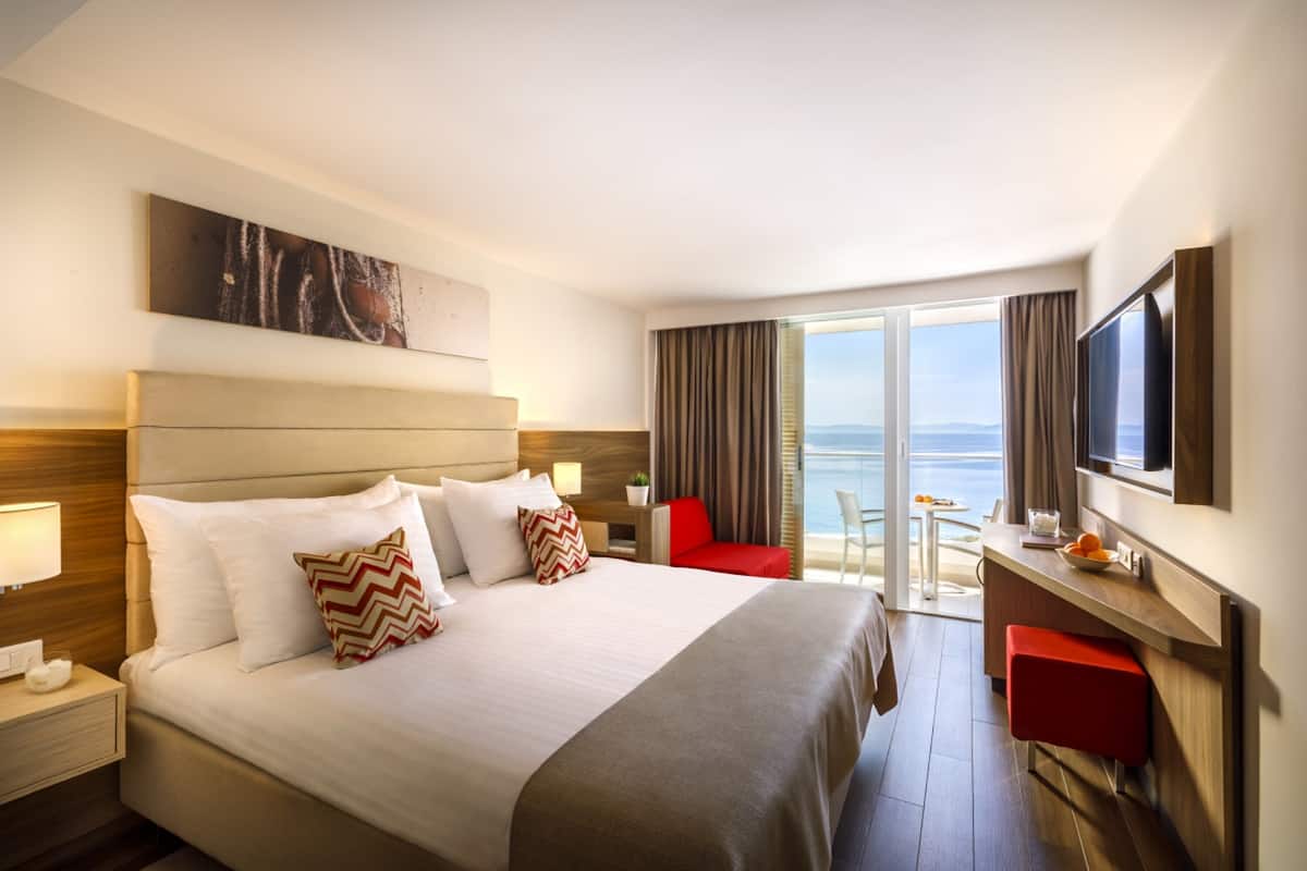A hotel room with a large bed, a desk with a chair, a TV, and a view of the sea.
