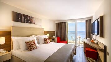 A hotel room with a large bed, a desk with a chair, a TV, and a view of the sea.