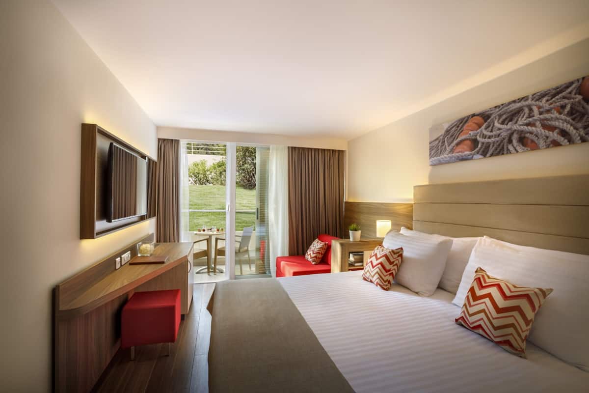 A hotel room with a bed, a desk, a TV, a red chair, and a sliding glass door to the outside.