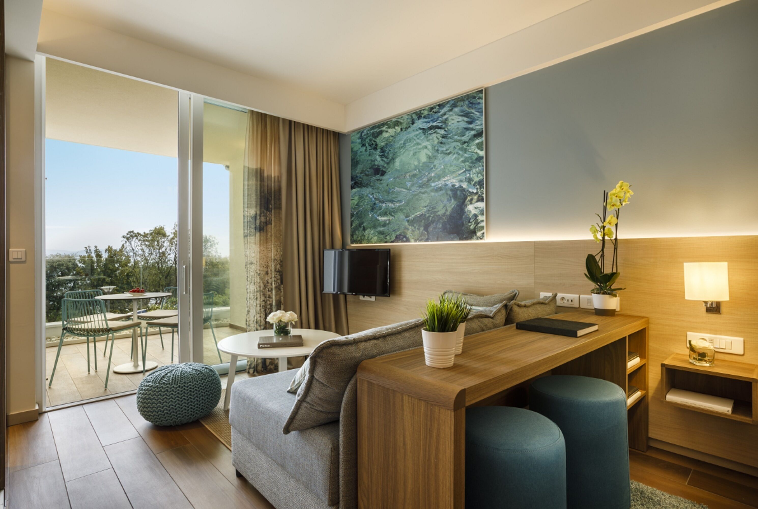 A modern hotel room with a sofa, a small dining table, a TV mounted on the wall, and a large window offering a view of the outdoors.