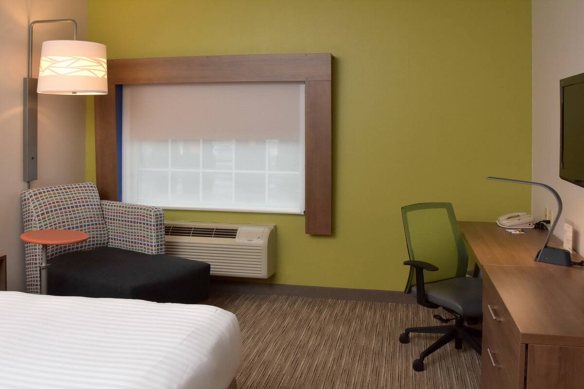 A hotel room with a bed, a desk, a chair, a lamp, a television, and a window with blinds.