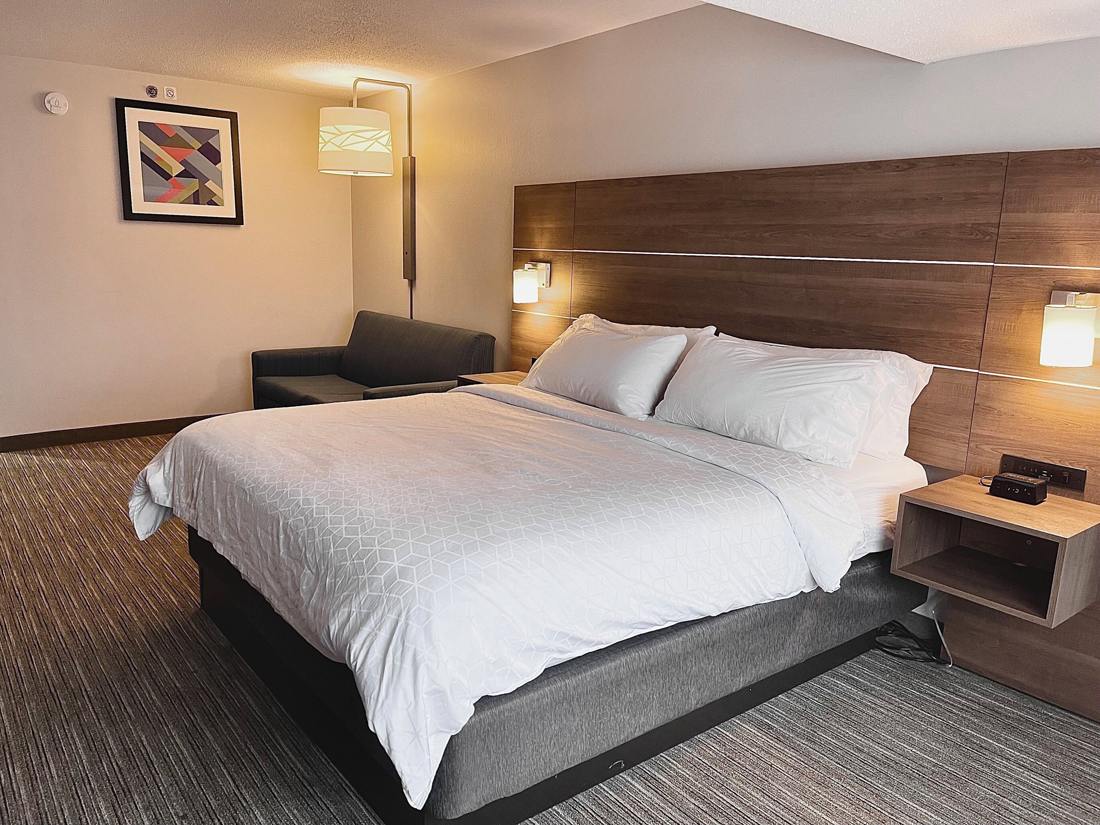 Quarto de hotel com uma cama, mesas de cabeceira, uma cadeira e uma luminária na parede.