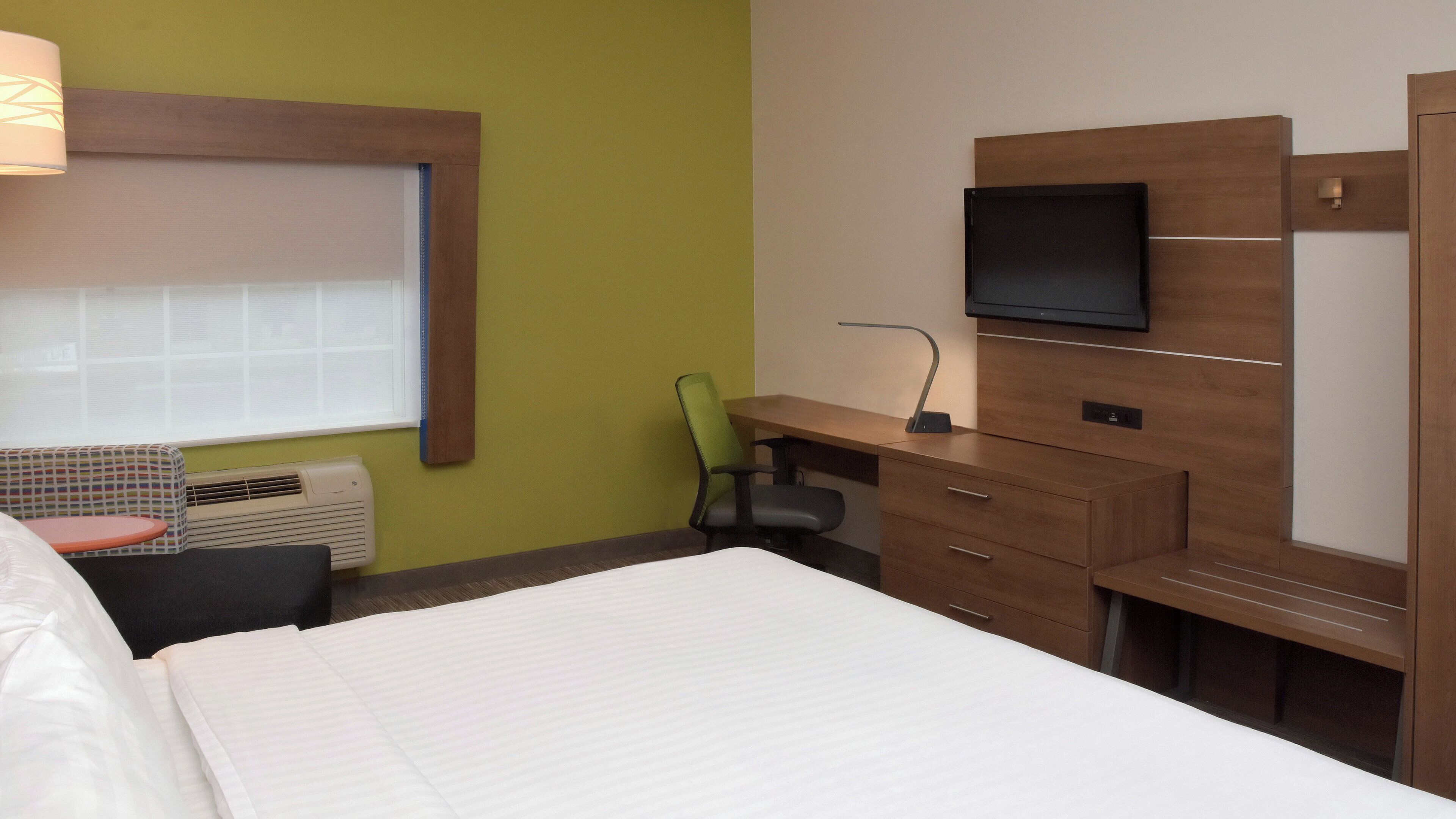 A hotel room with a bed, a desk with a chair, a television, and a window with blinds.