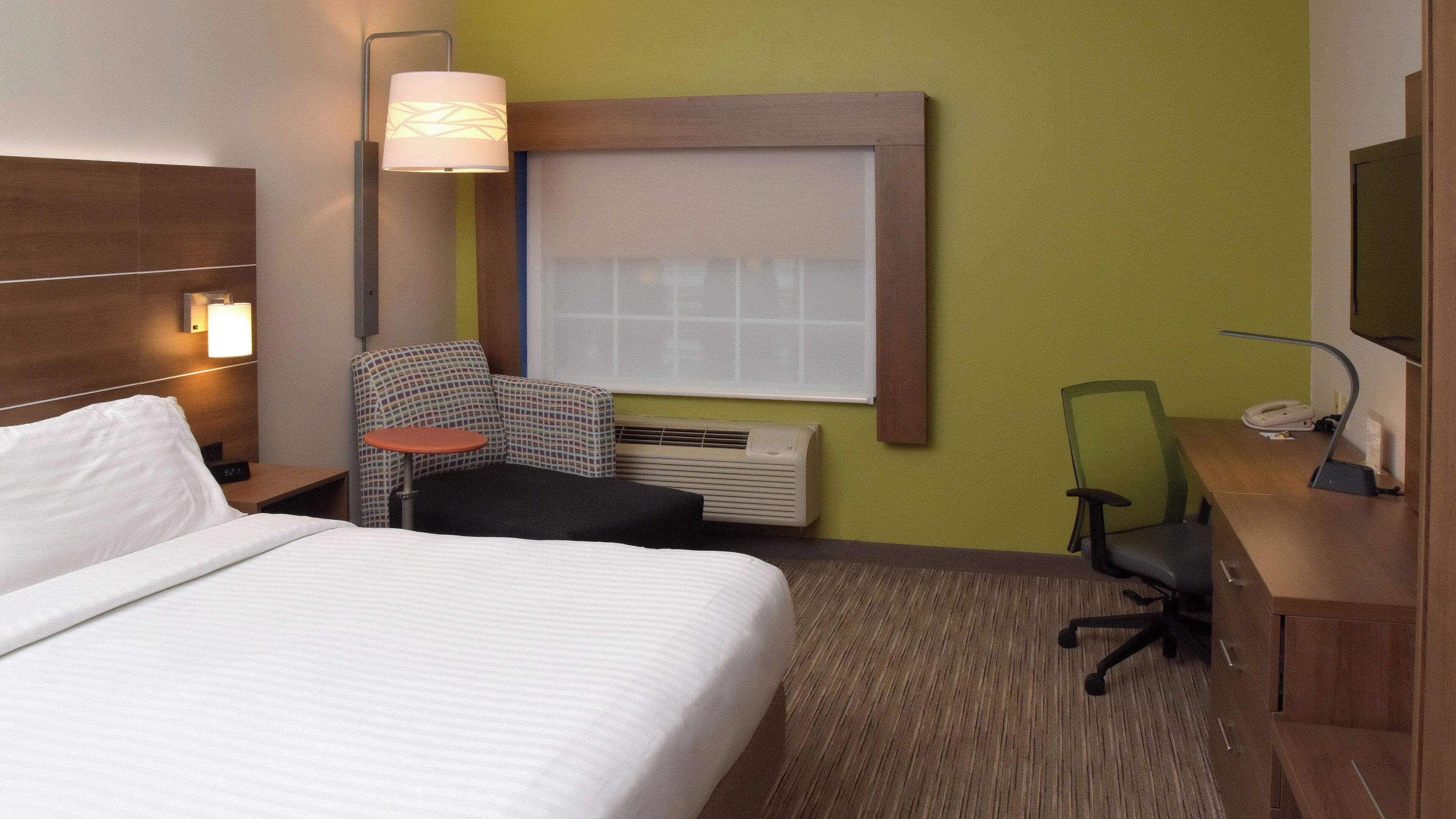 Une chambre d’hôtel comprenant un lit, un bureau, une chaise, une petite table, une lampe, un radiateur et une fenêtre.