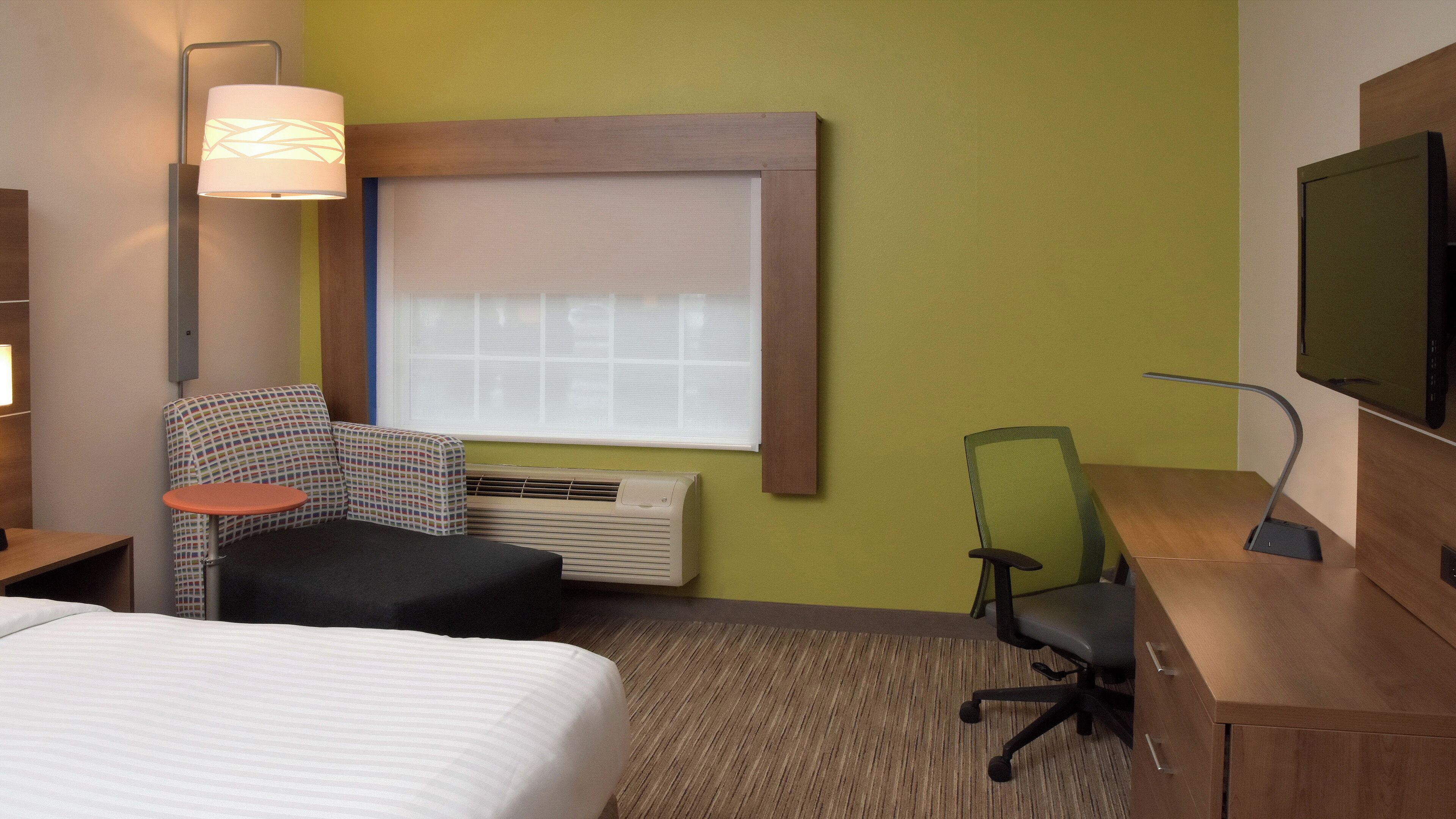 A hotel room with a bed, a desk, a chair, a TV, a window with blinds, and a lamp.