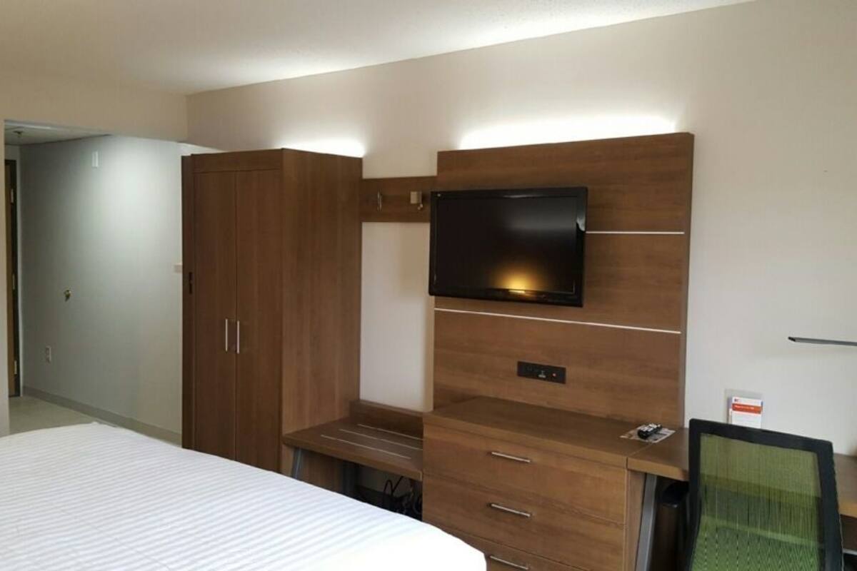 A hotel room with a bed, a TV mounted on a wooden cabinet, a desk with a chair, and a wardrobe.