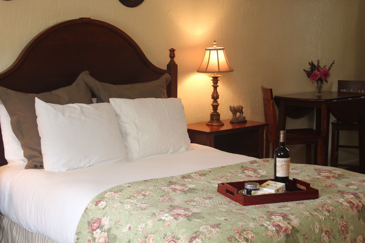 A neatly made bed with white linens, a floral bedspread, a bedside lamp, a bottle of wine, and a tray with a candle and a box on the bed.