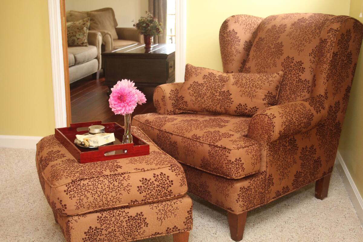 A living room with a patterned armchair and ottoman, a small table with a vase of flowers, and a tray with candles and a book.
