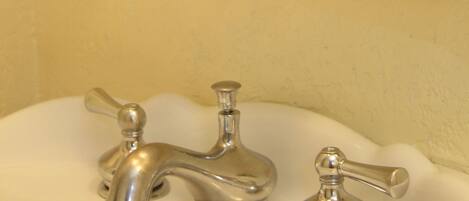 A bathroom with a glass shelf holding a potted plant, a bottle of lotion, and a rolled towel. There are two faucets on the sink.