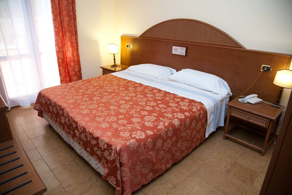 A hotel room with a large bed, two bedside tables with lamps, a telephone, and a window with curtains.