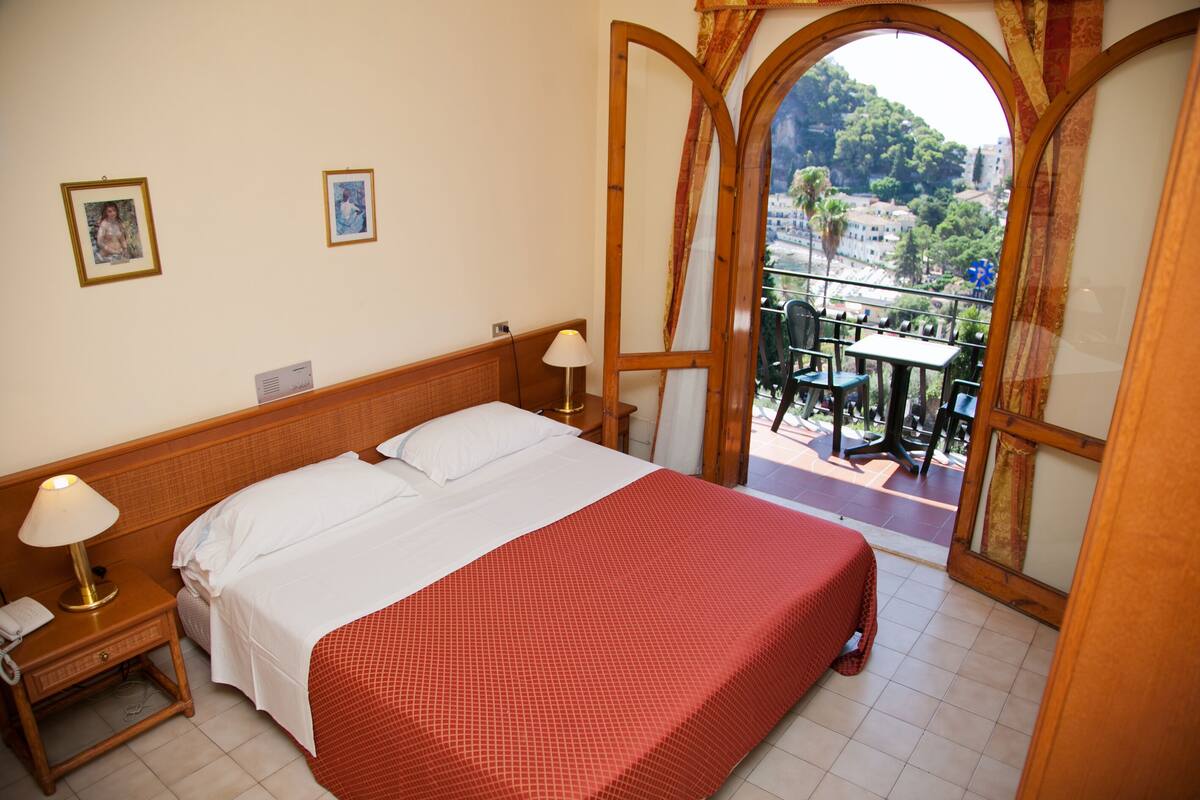 A hotel room with a bed, nightstand, lamp, and a balcony with a table and chairs.