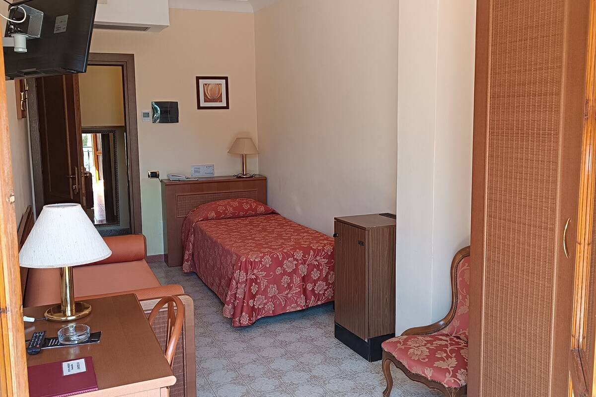 A hotel room with a bed, a desk with a lamp, a chair, a wardrobe, and a television.