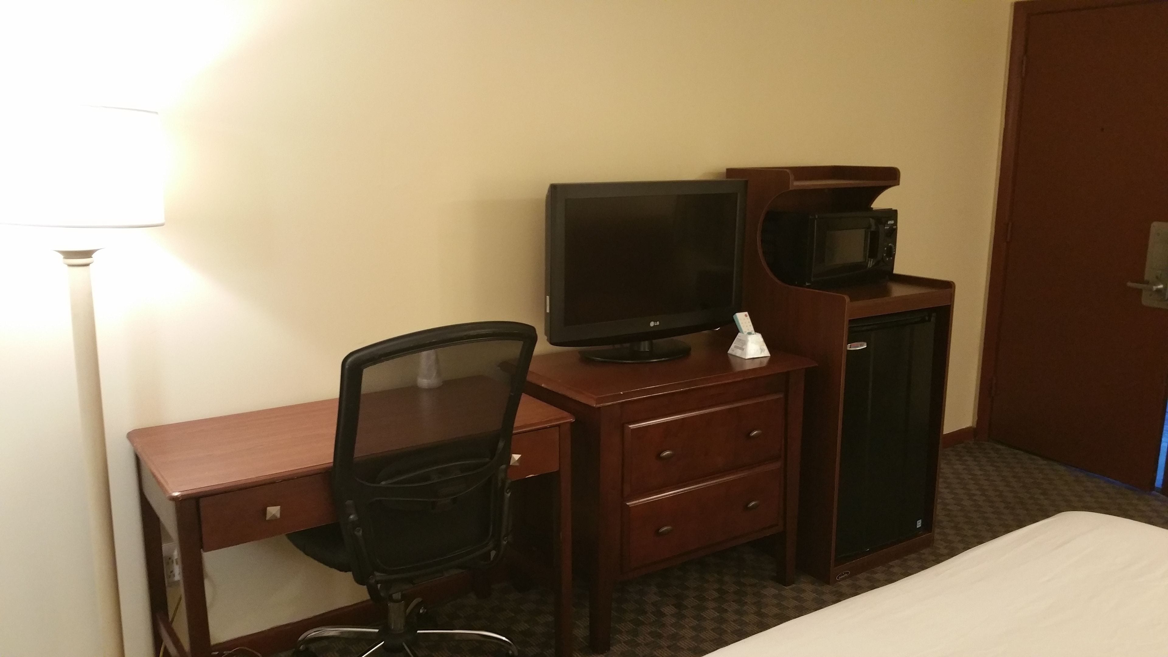 A hotel room with a wooden desk, a black office chair, a flat-screen TV, a microwave, and a small cabinet.