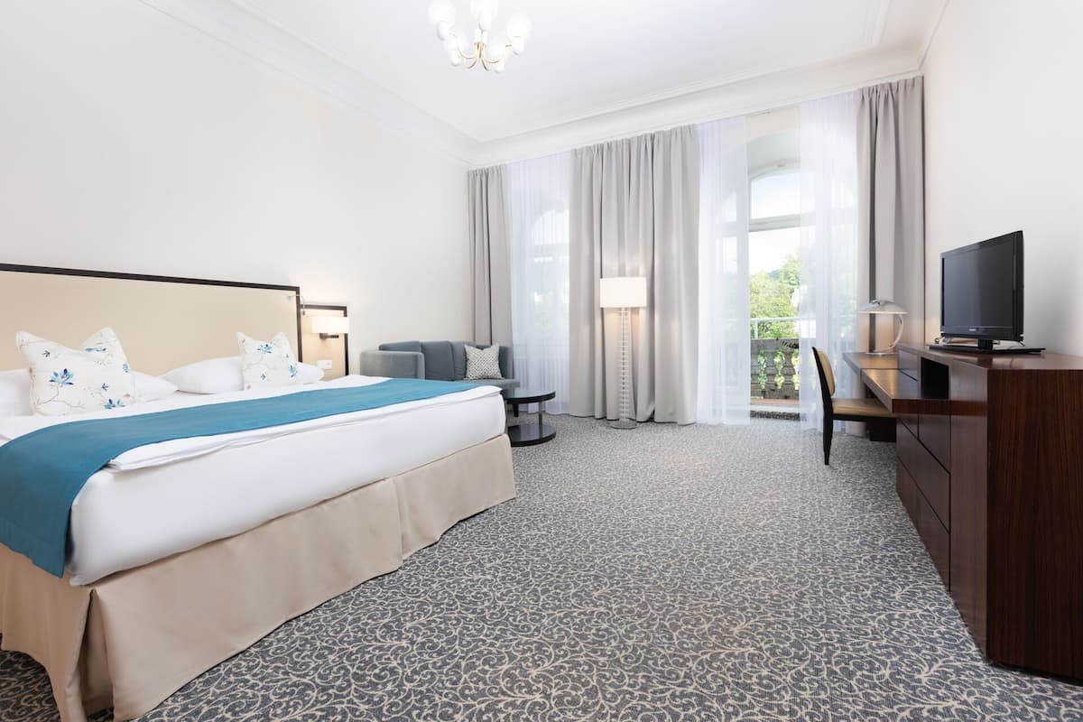Ein Hotelzimmer mit einem großen Bett, einem Schreibtisch mit Fernseher, einem Sessel und Blick ins Grüne.
