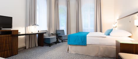 A hotel room with a bed, a desk with a chair, a TV, and a window with curtains.