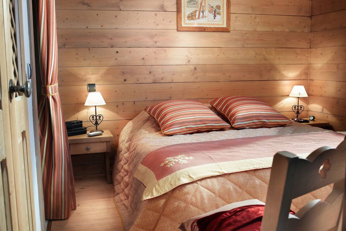 Una habitación de madera con una cama, dos mesitas de noche, una silla y una puerta.