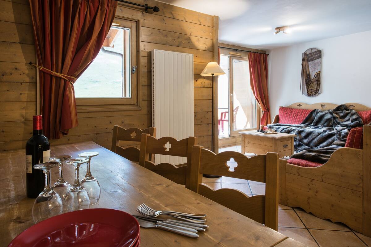 Un interior de madera con una mesa de comedor, sillas, una cama y un ventanal con cortinas.