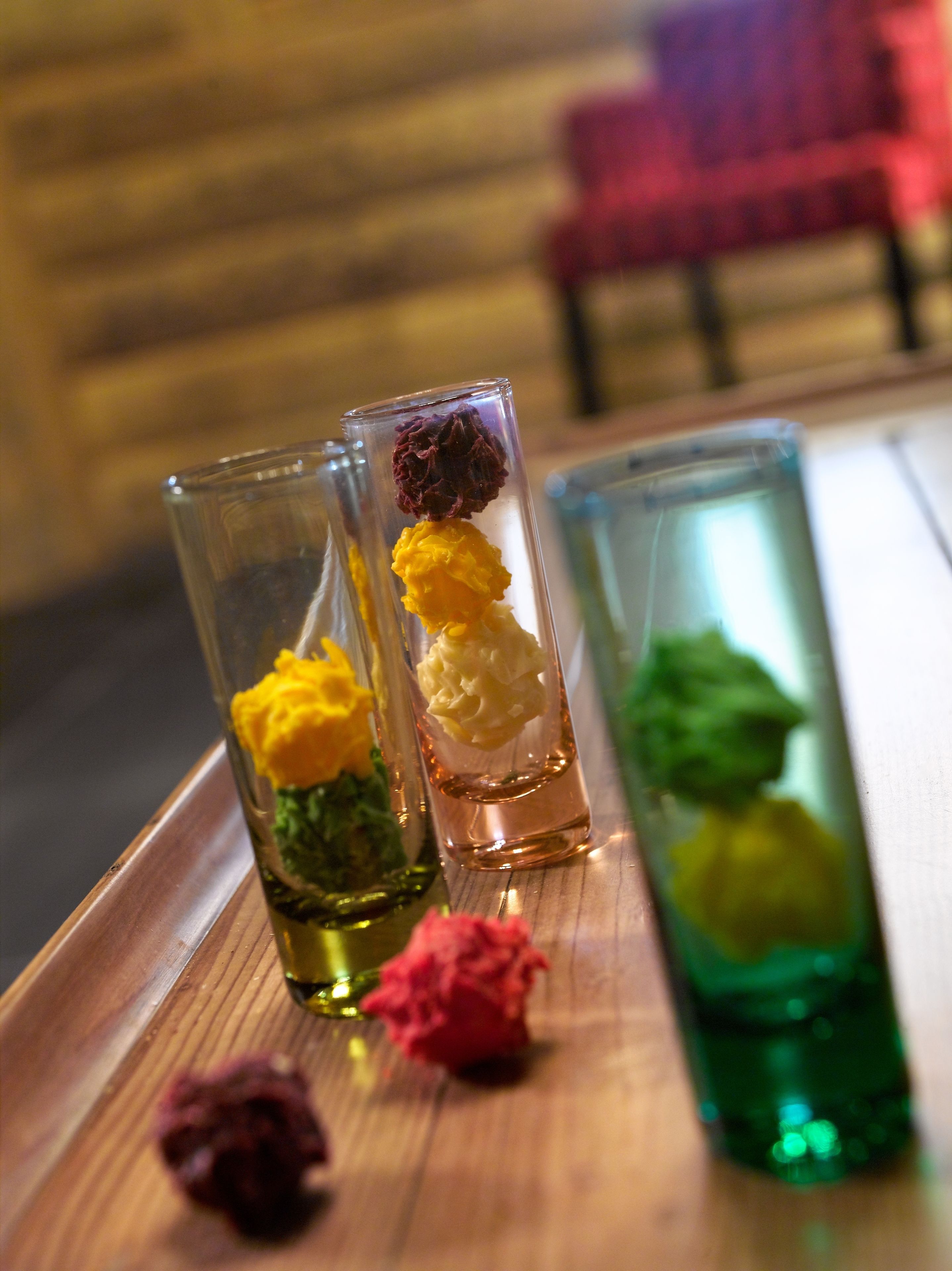 A wooden table with three clear shot glasses containing colorful flower arrangements.