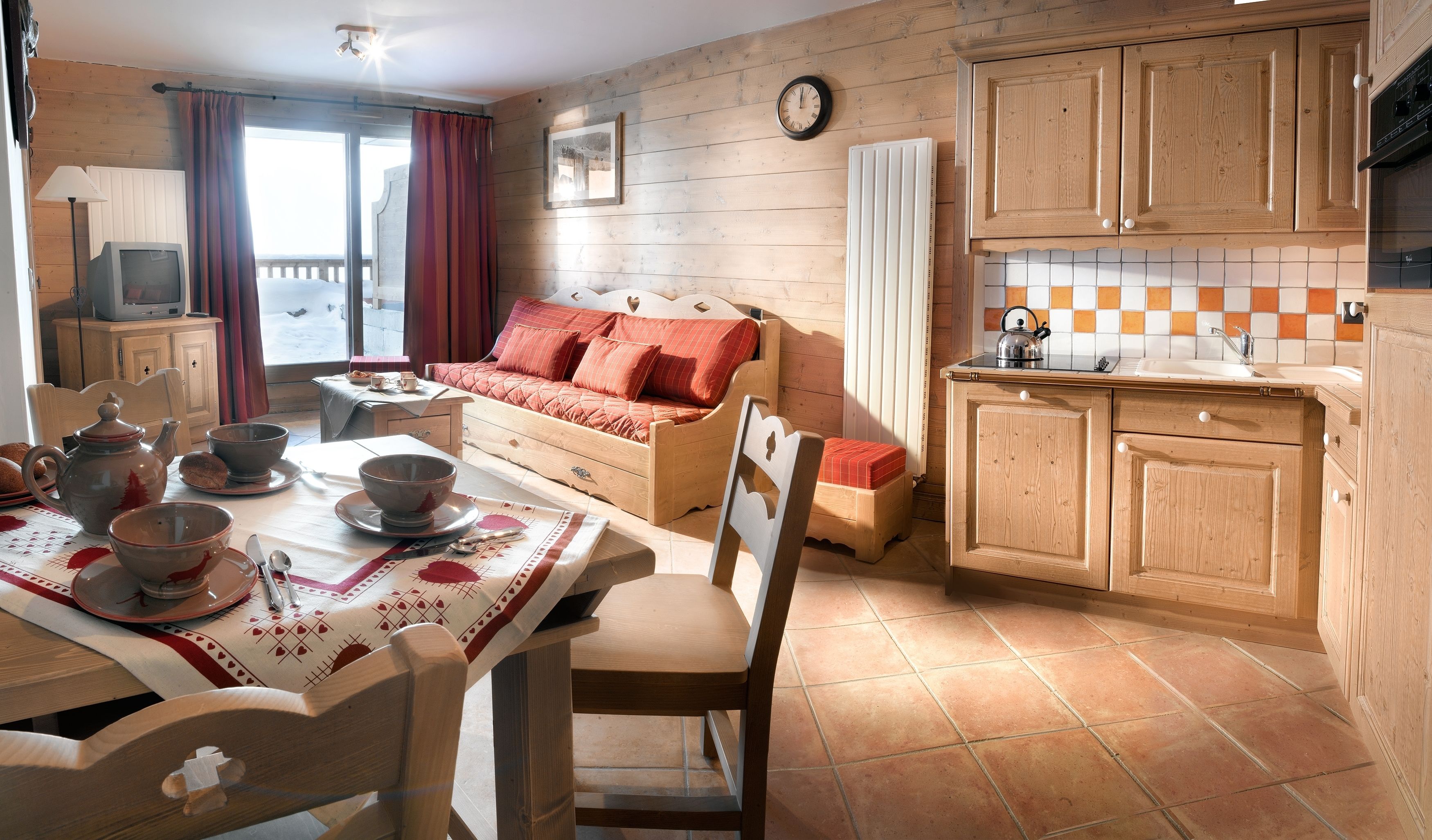 Une pièce chaleureuse avec un canapé en bois, une table à manger recouverte d’une nappe rouge et blanche, et une cuisine dotée de meubles en bois et d’un dosseret carrelé.