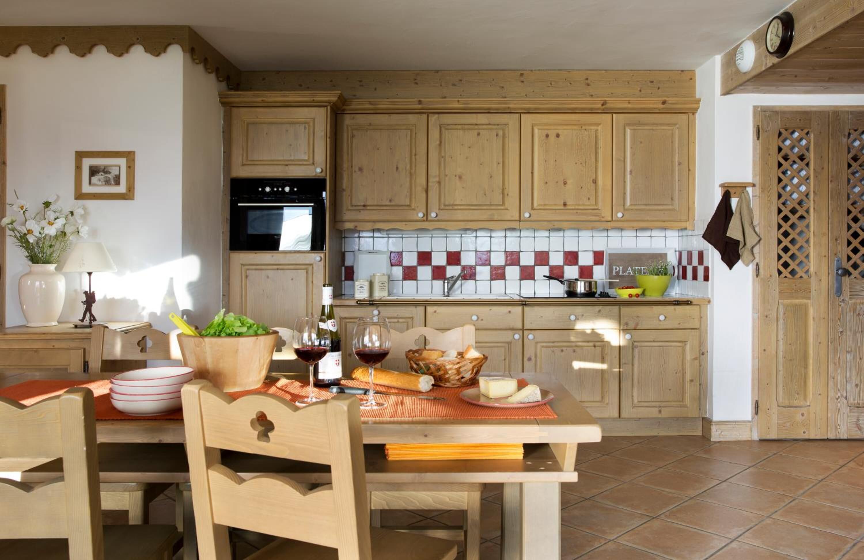 A kitchen with wooden cabinets, a tiled backsplash, and a dining table set with food and wine.