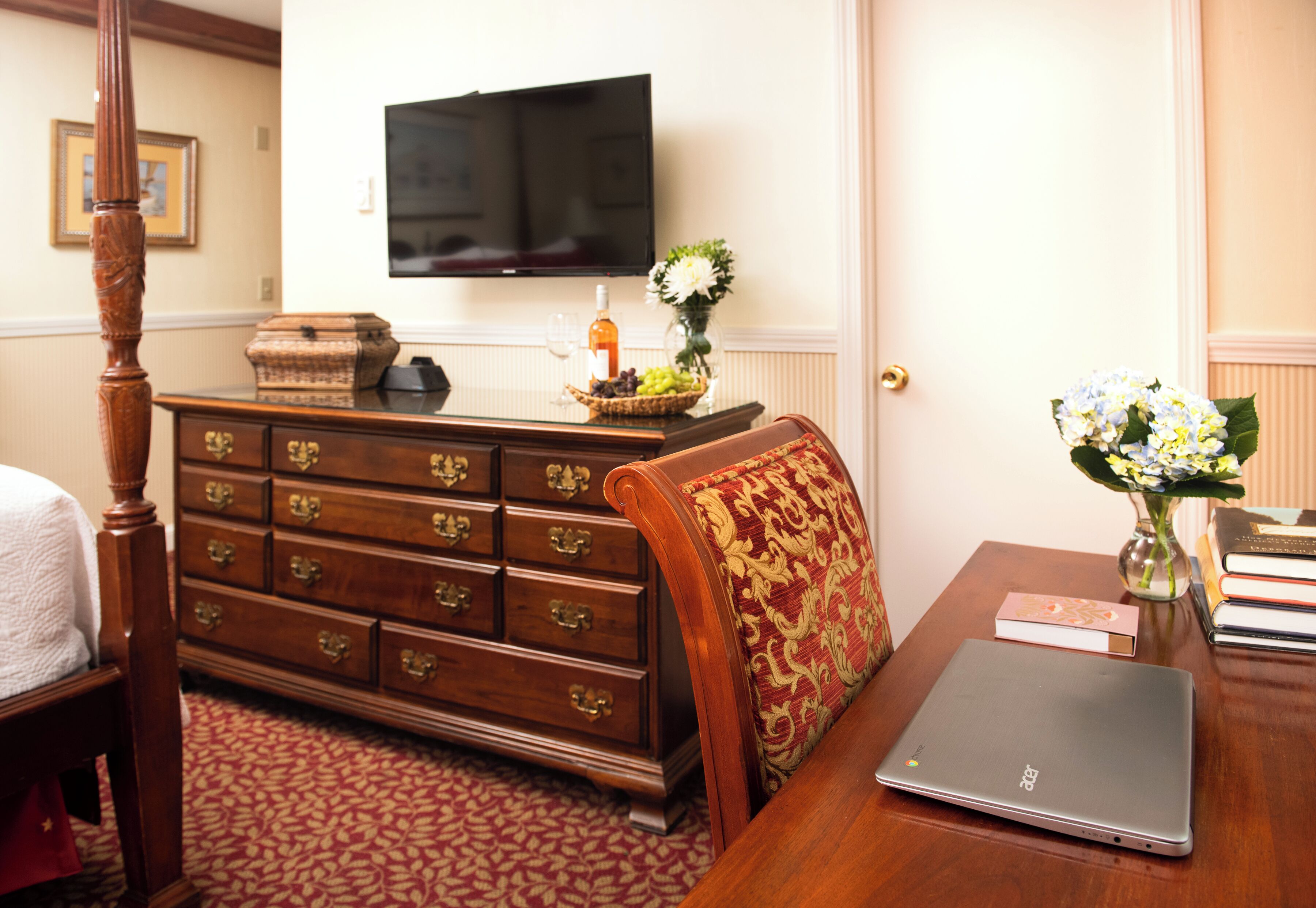 A hotel room with a wooden dresser, a laptop on a table, a television on the wall, and a vase with flowers.