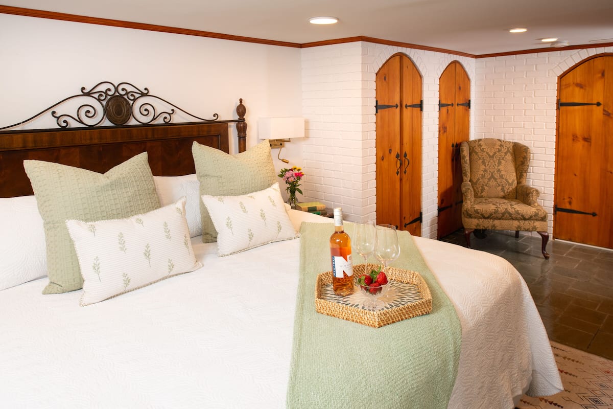 A neatly made bed with pillows, a bedside table with a bottle and glasses, and a chair in a room with white brick walls and wooden doors.