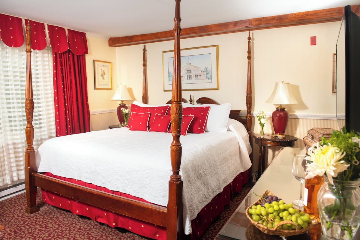 A four-poster bed with red bedding, a vase of flowers, and a basket of fruit on a bedside table.