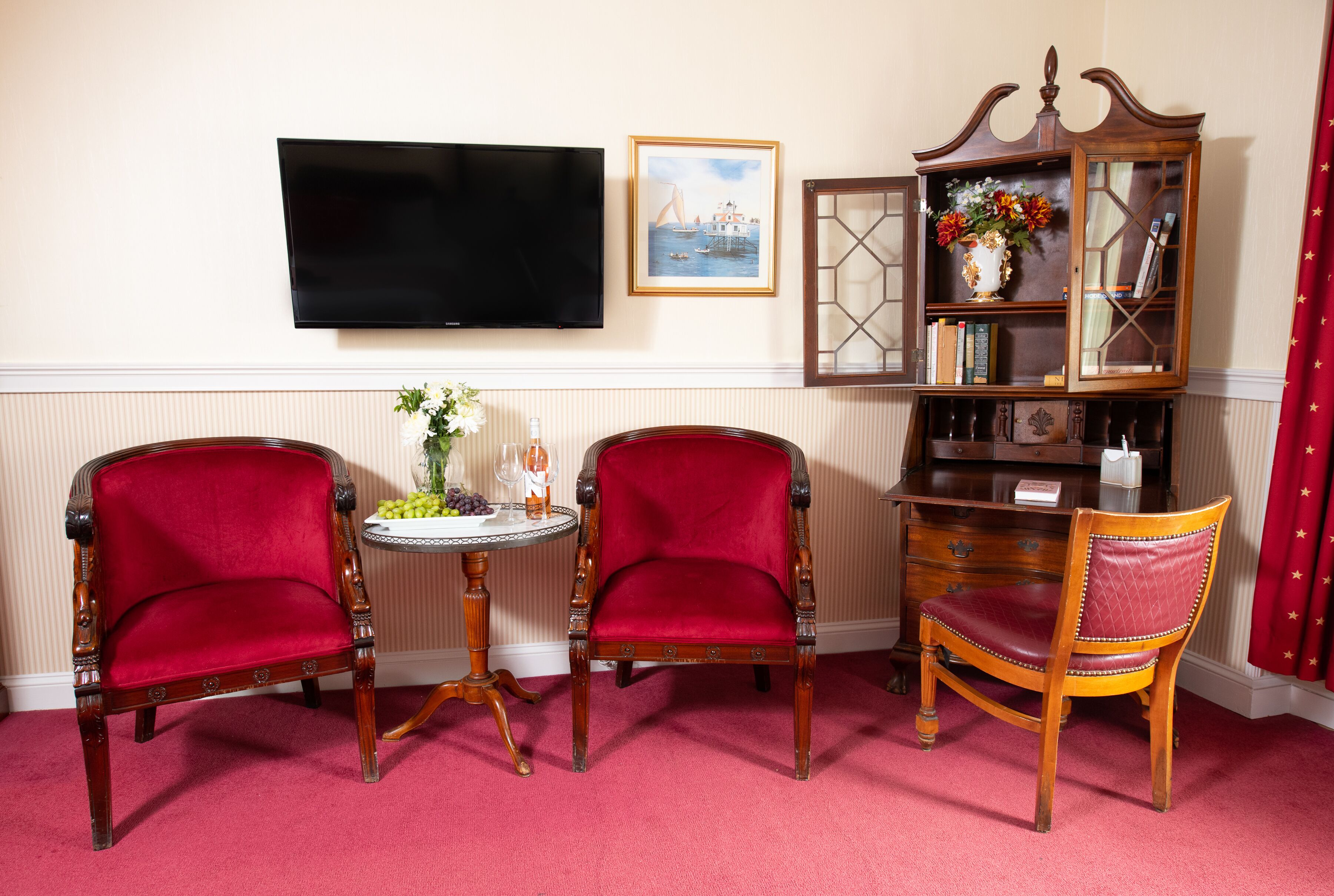 Ein Zimmer mit zwei roten Polsterstühlen, einem kleinen runden Tisch mit einer Vase und Obst, einem Holzregal und einem an der Wand befestigten Flachbildfernseher.